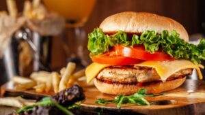 American cuisine concept. Juicy burger with meat patty, tomatoes, cheddar cheese, lettuce and homemade bun. In the background are french fries and a glass of beer. Close up