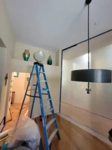 A room under renovation featuring a ladder, decorative vases, and a large pendant light. Walls are taped for painting.